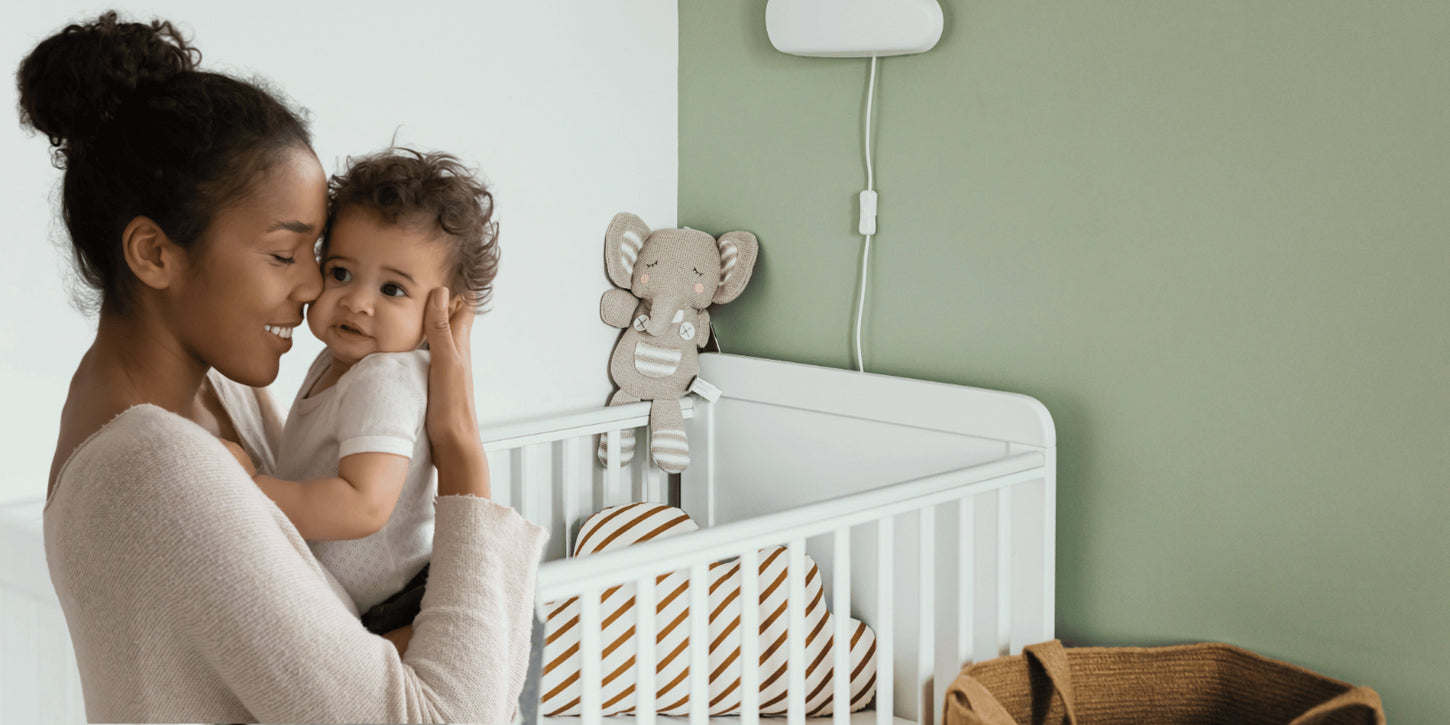 Smiling mother holding her baby next to a white crib with a plush elephant toy and striped pillows.