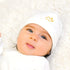 Newborn baby wearing a white outfit and hat with a yellow cloud and stars design, lying calmly with eyes open.