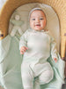 Smiling baby in a mint green striped romper and bonnet from Calm Nest, lying in a cozy bassinet with a soft toy.